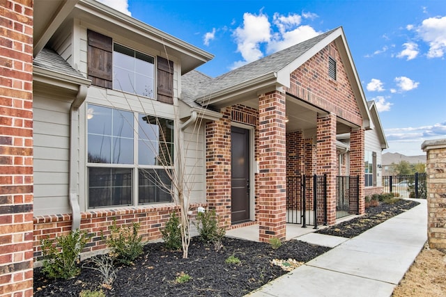 view of exterior entry featuring brick siding, a shingled roof, and fence