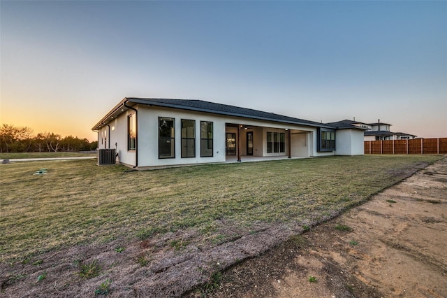 back of house at dusk featuring central air condition unit, fence, a lawn, stucco siding, and a patio area