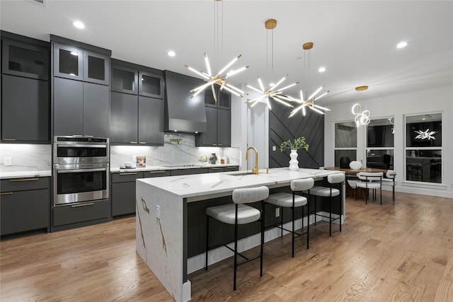 kitchen with glass insert cabinets, hanging light fixtures, stainless steel double oven, a large island with sink, and a sink