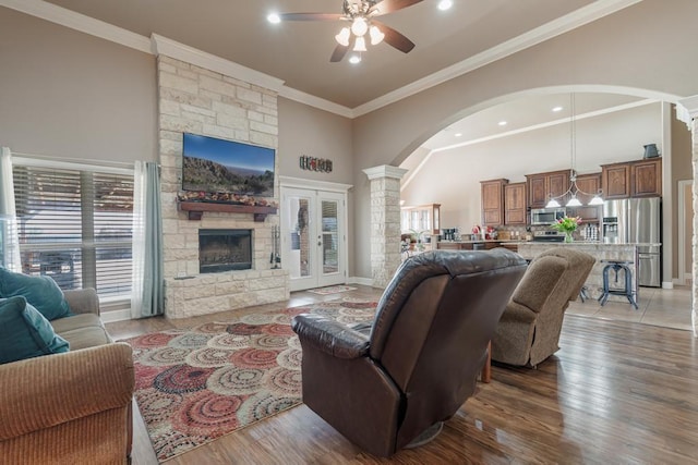 living room with arched walkways, a towering ceiling, wood finished floors, crown molding, and a stone fireplace