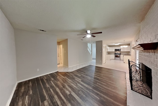 unfurnished living room featuring a textured ceiling, ceiling fan with notable chandelier, wood finished floors, visible vents, and a brick fireplace