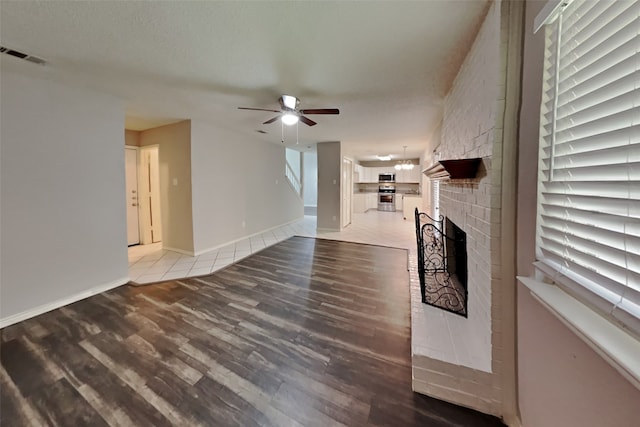 unfurnished living room with ceiling fan with notable chandelier, wood finished floors, visible vents, baseboards, and a brick fireplace