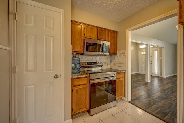 kitchen with stainless steel appliances, backsplash, stone countertops, brown cabinetry, and a textured ceiling