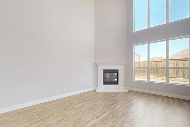 unfurnished living room featuring light wood-type flooring, a glass covered fireplace, a towering ceiling, and baseboards