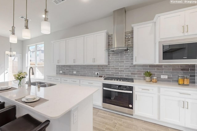 kitchen with stainless steel appliances, light countertops, hanging light fixtures, white cabinetry, and wall chimney range hood