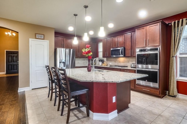 kitchen with pendant lighting, stainless steel appliances, tasteful backsplash, a kitchen island with sink, and light stone countertops