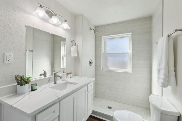 bathroom with toilet, a textured wall, a tile shower, and vanity