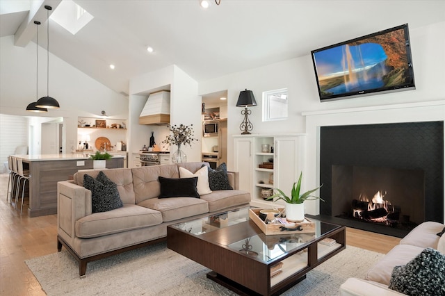 living area featuring a warm lit fireplace, high vaulted ceiling, a skylight, light wood finished floors, and beamed ceiling