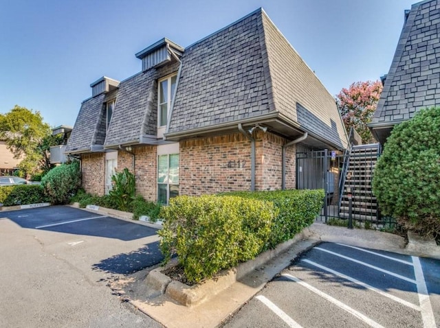 view of property exterior featuring a shingled roof, mansard roof, stairs, uncovered parking, and brick siding