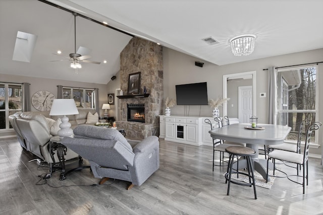 living room featuring light wood-type flooring, ceiling fan with notable chandelier, visible vents, and a stone fireplace