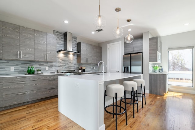 kitchen with a center island with sink, stainless steel fridge with ice dispenser, wall chimney exhaust hood, modern cabinets, and light countertops