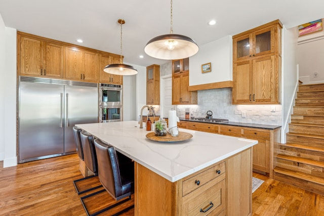 kitchen featuring brown cabinetry, an island with sink, glass insert cabinets, decorative light fixtures, and stainless steel appliances