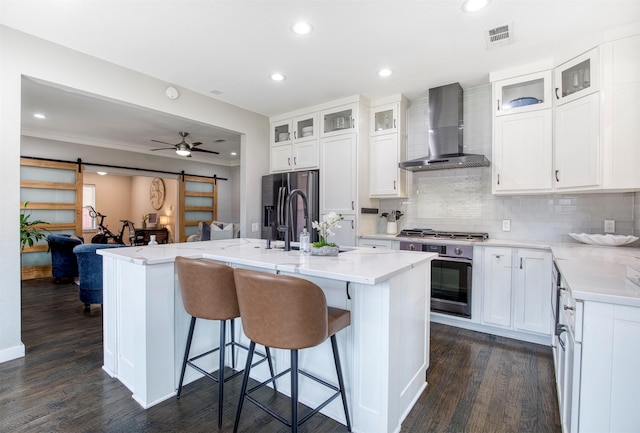 kitchen with a center island with sink, a barn door, appliances with stainless steel finishes, glass insert cabinets, and wall chimney exhaust hood