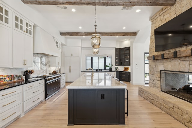 kitchen with custom range hood, double oven range, an island with sink, glass insert cabinets, and pendant lighting