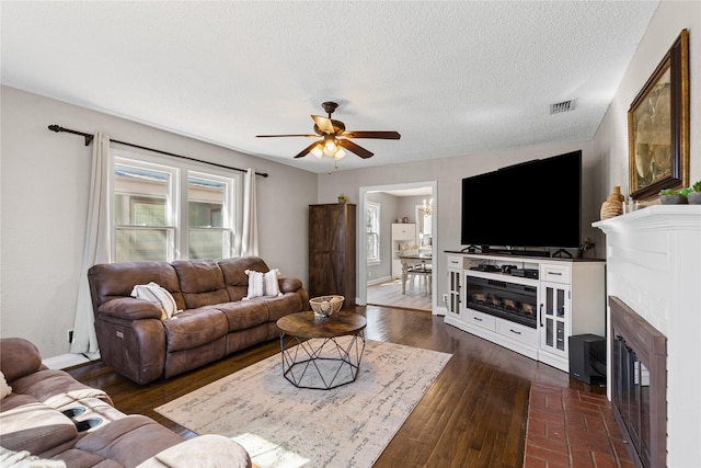 living room featuring dark wood-style floors, visible vents, a ceiling fan, a glass covered fireplace, and a textured ceiling