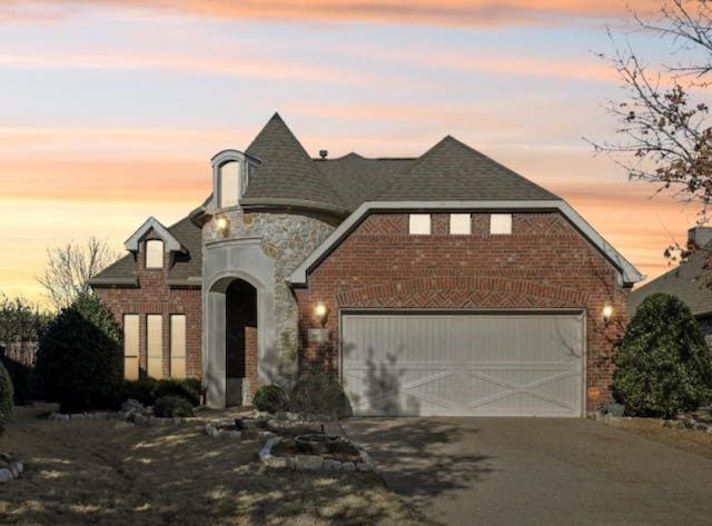 french country style house with a garage, driveway, brick siding, and stone siding