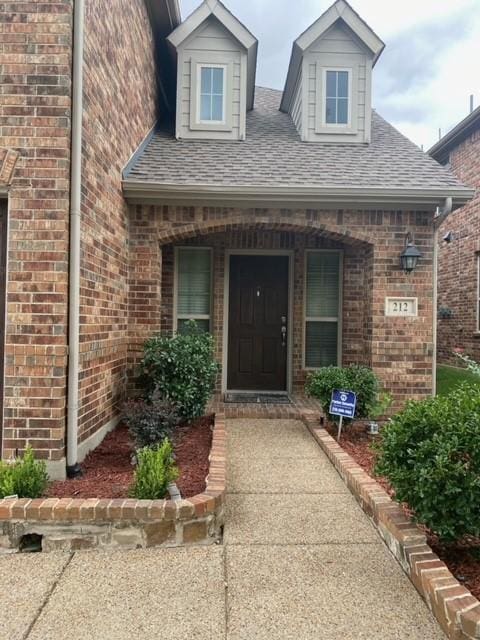 doorway to property with covered porch, brick siding, and roof with shingles