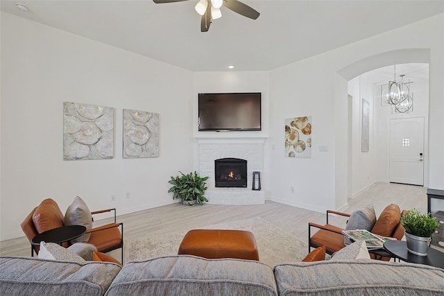 living room with a warm lit fireplace, baseboards, arched walkways, light wood-style floors, and ceiling fan with notable chandelier