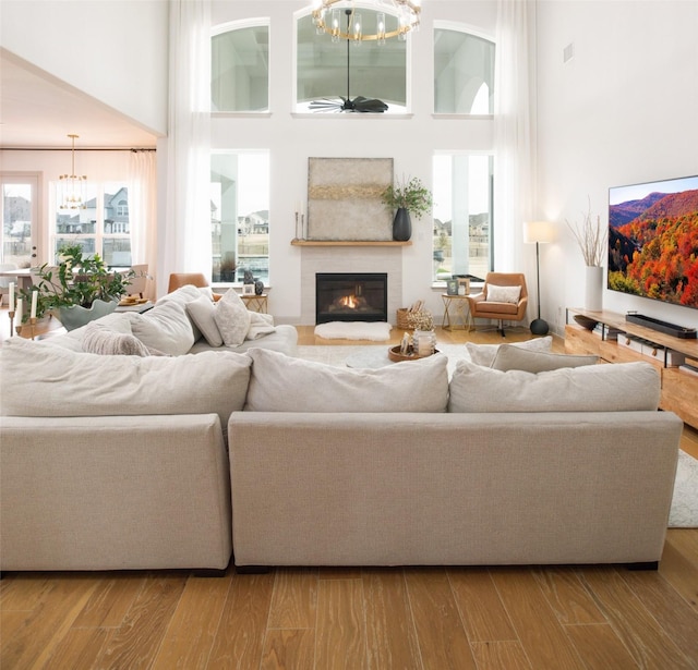 living room featuring wood finished floors, a glass covered fireplace, a towering ceiling, and ceiling fan with notable chandelier