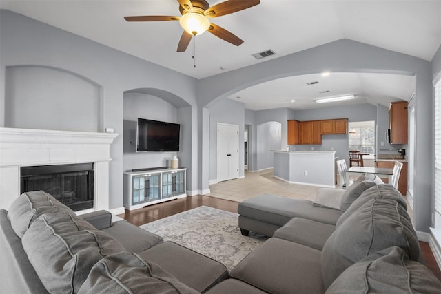 living room with a fireplace with raised hearth, ceiling fan, visible vents, baseboards, and vaulted ceiling