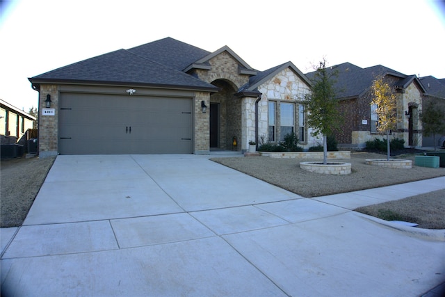 view of front of property featuring roof with shingles, concrete driveway, central AC, a garage, and stone siding