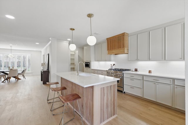 kitchen with a kitchen island with sink, a sink, light wood-style floors, hanging light fixtures, and high end stainless steel range