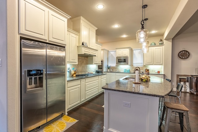 kitchen featuring a center island with sink, appliances with stainless steel finishes, white cabinetry, a sink, and dark stone counters