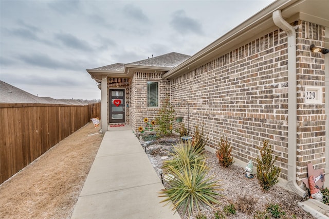 doorway to property featuring brick siding, fence, and roof with shingles