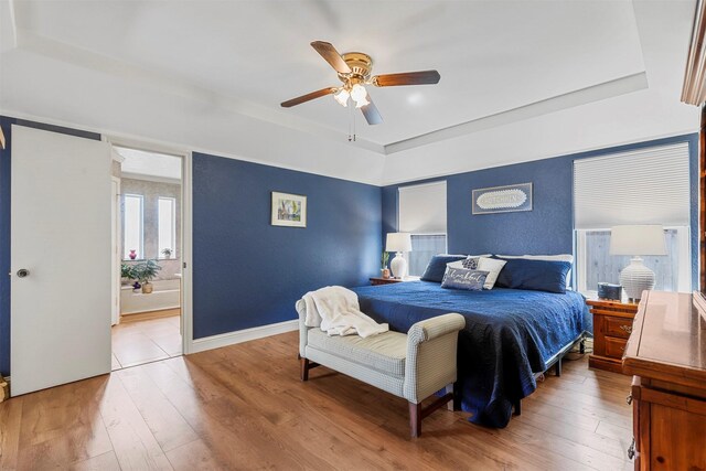 bedroom featuring baseboards, a tray ceiling, ceiling fan, light wood-type flooring, and connected bathroom