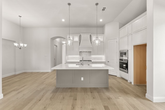 kitchen featuring wall oven, a kitchen island with sink, premium range hood, light countertops, and stainless steel microwave