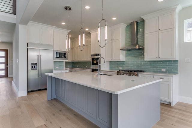 kitchen featuring a center island with sink, hanging light fixtures, white cabinetry, built in appliances, and wall chimney exhaust hood