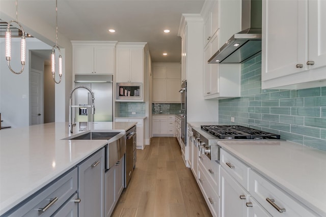 kitchen with white cabinets, built in appliances, hanging light fixtures, light countertops, and wall chimney range hood