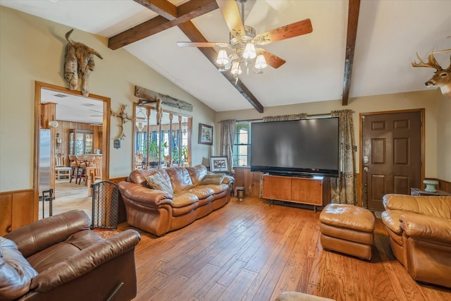 living area featuring lofted ceiling with beams, a ceiling fan, wainscoting, wood walls, and light wood-type flooring