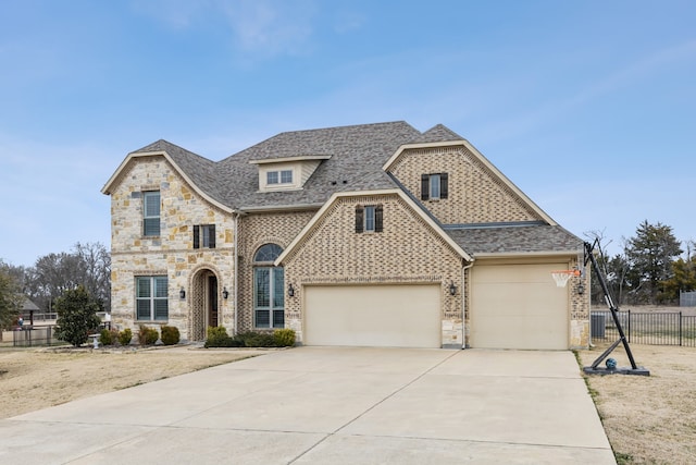 french country home featuring a shingled roof, concrete driveway, stone siding, an attached garage, and fence