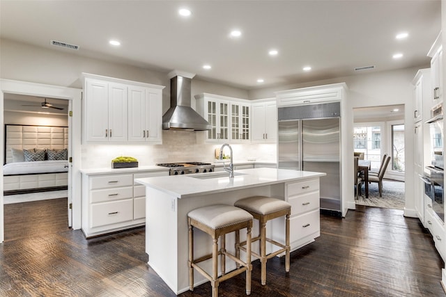 kitchen with a sink, wall chimney range hood, stainless steel built in fridge, an island with sink, and glass insert cabinets