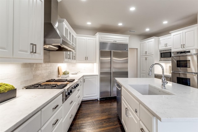 kitchen with white cabinets, built in appliances, light stone countertops, wall chimney range hood, and a sink