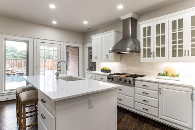 kitchen featuring stainless steel gas cooktop, a sink, white cabinetry, wall chimney exhaust hood, and a center island with sink