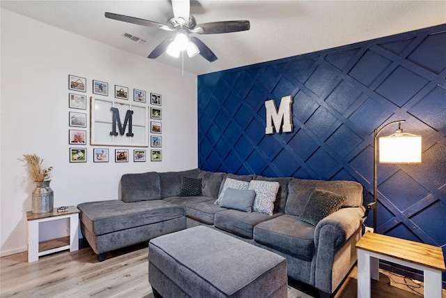 living room featuring an accent wall, wood finished floors, visible vents, baseboards, and a ceiling fan