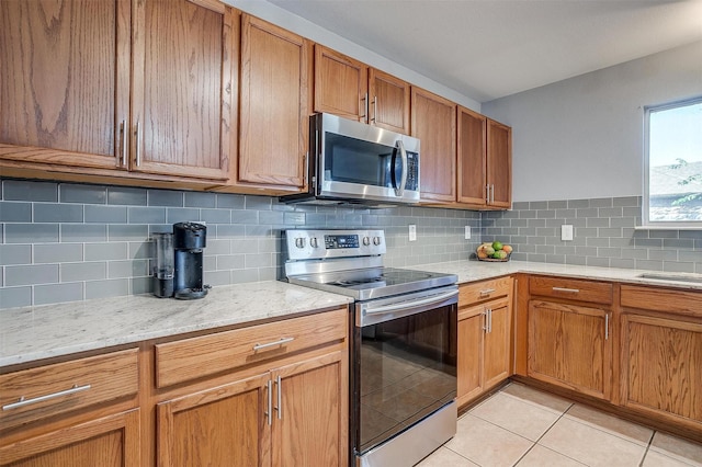 kitchen featuring brown cabinetry, light stone countertops, light tile patterned flooring, stainless steel appliances, and backsplash