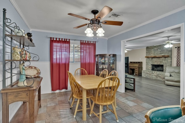 dining room featuring visible vents, ornamental molding, a ceiling fan, a brick fireplace, and wainscoting