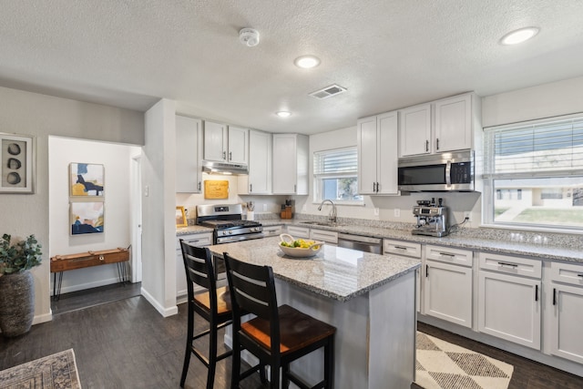 kitchen with under cabinet range hood, stainless steel appliances, a kitchen island, white cabinets, and light stone countertops