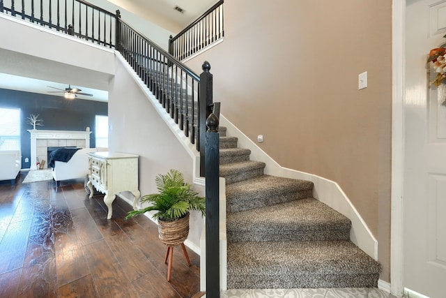 stairs featuring ceiling fan, a fireplace, a towering ceiling, visible vents, and hardwood / wood-style floors