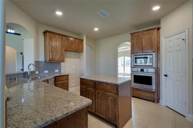 kitchen featuring stainless steel appliances, a sink, a center island, light stone countertops, and brown cabinetry