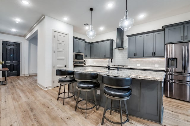 kitchen featuring a center island with sink, stainless steel appliances, a sink, light stone countertops, and wall chimney exhaust hood