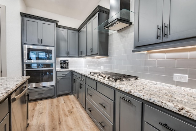 kitchen with gray cabinetry, appliances with stainless steel finishes, light wood-type flooring, backsplash, and wall chimney exhaust hood