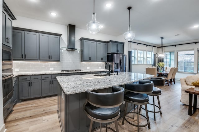 kitchen with wall chimney exhaust hood, a kitchen island with sink, stainless steel appliances, and decorative light fixtures