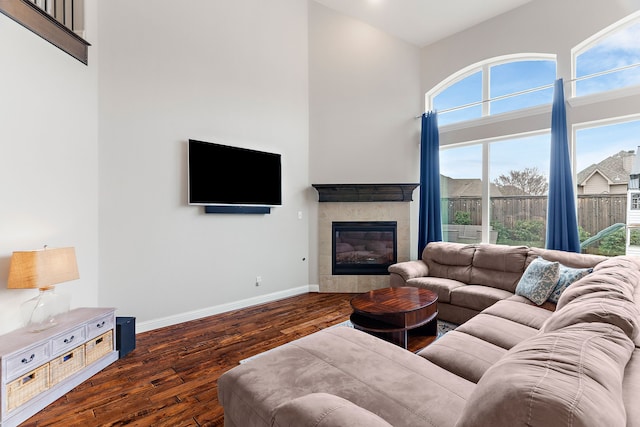 living area with a high ceiling, a tiled fireplace, dark wood finished floors, and baseboards