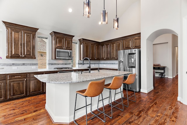 kitchen with arched walkways, stainless steel appliances, a sink, light stone countertops, and a center island with sink