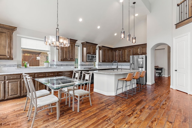 kitchen featuring light stone counters, arched walkways, pendant lighting, stainless steel appliances, and a kitchen island with sink