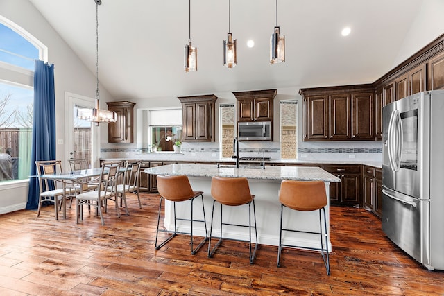 kitchen with an island with sink, lofted ceiling, appliances with stainless steel finishes, hanging light fixtures, and light stone countertops